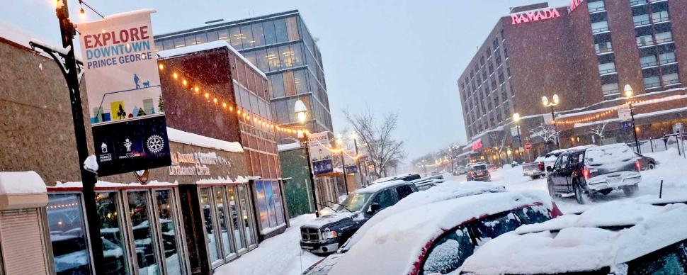 Cars parked in snow in downtown Prince George during a winter evening.