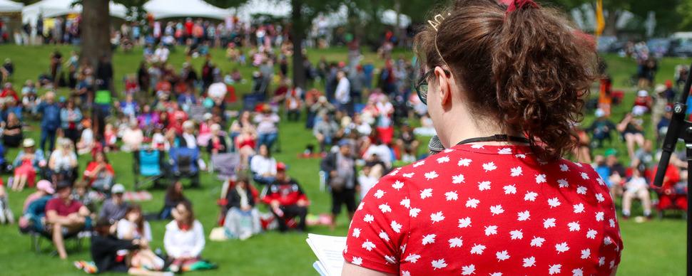 The back of a person on stage addressing a crowd on a grassy field.