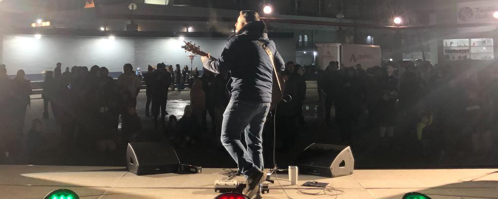 A performer with an acoustic guitar on stage at night, with a silhouetted crowd in the background.
