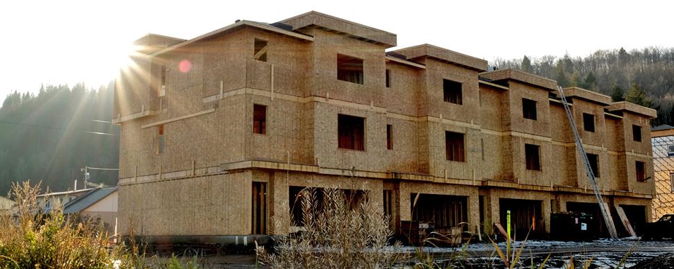 Unfinished exterior of a 5 unit townhouse complex. Grass in foreground. Sun setting behind tree-covered hill in background.